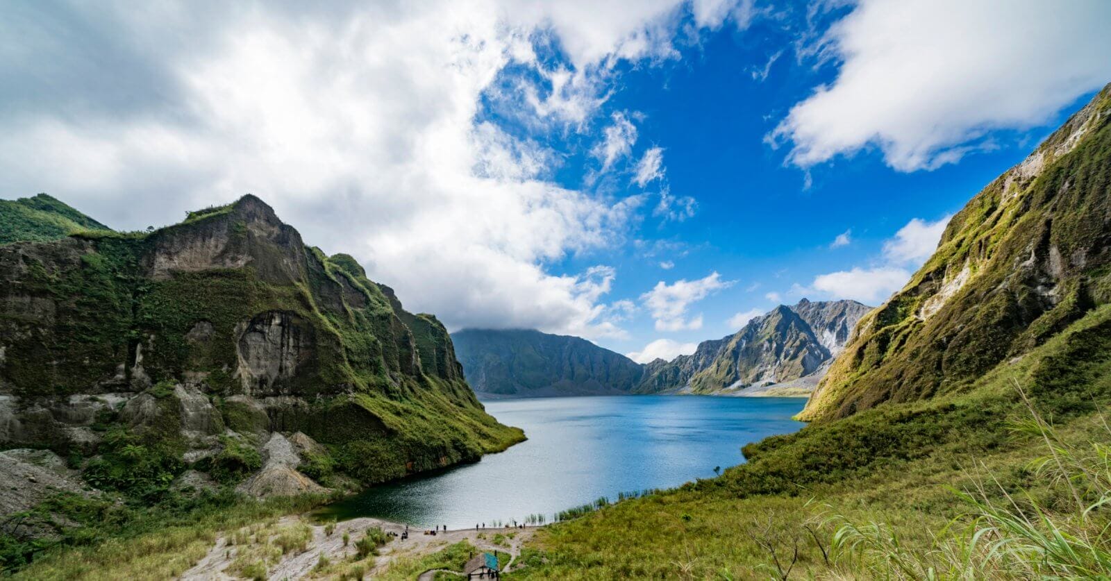 A pristine lake is nestled between towering, green-covered mountains in Region 3 under a partly cloudy sky. The vibrant blue water reflects the surrounding cliffs and the azure of the sky. Lush vegetation covers the mountainsides, and a few people are visible on a path near the lake.