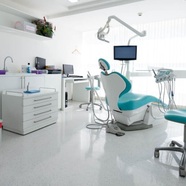A modern dental office in Iligan City with white walls and floors. The room features a dental chair with an overhead light, a monitor, and various instruments. Two additional blue and white chairs are present. Cabinets line the left wall, and a desk with a computer is in the background — one of the best dental clinics around.