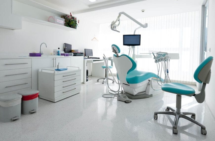 A modern dental office in Iligan City with white walls and floors. The room features a dental chair with an overhead light, a monitor, and various instruments. Two additional blue and white chairs are present. Cabinets line the left wall, and a desk with a computer is in the background — one of the best dental clinics around.