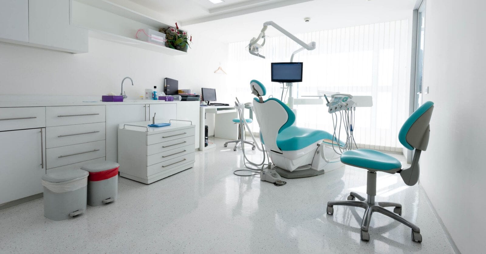 A modern dental office in Iligan City with white walls and floors. The room features a dental chair with an overhead light, a monitor, and various instruments. Two additional blue and white chairs are present. Cabinets line the left wall, and a desk with a computer is in the background — one of the best dental clinics around.