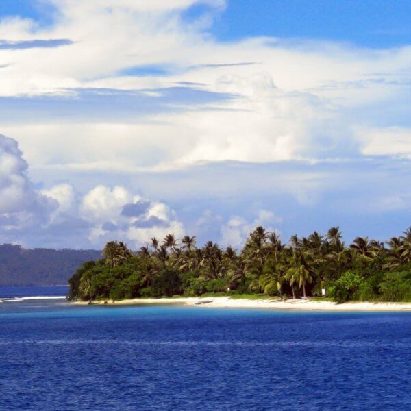 A picturesque tropical island in Eastern Visayas is surrounded by blue ocean water. The coastline, within Region 8's stunning locales, is lined with lush green palm trees and greenery that stands out against the sandy beach. In the background, hills and a partly cloudy sky with scattered white clouds meet the horizon.