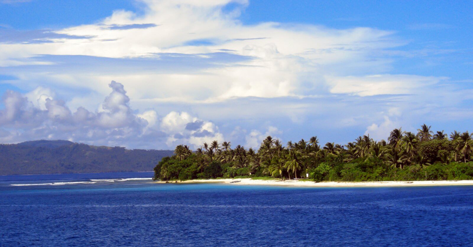A picturesque tropical island in Eastern Visayas is surrounded by blue ocean water. The coastline, within Region 8's stunning locales, is lined with lush green palm trees and greenery that stands out against the sandy beach. In the background, hills and a partly cloudy sky with scattered white clouds meet the horizon.