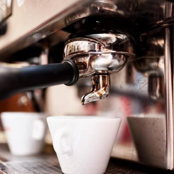 Close-up of a professional espresso machine brewing coffee into a white ceramic cup. The portafilter and spout are shiny and metallic, fixed securely in place. The cup sits on a metal drip tray, catching dark espresso as it flows from the machine. Experience this at one of the best restaurants in Gateway Mall, Quezon City.