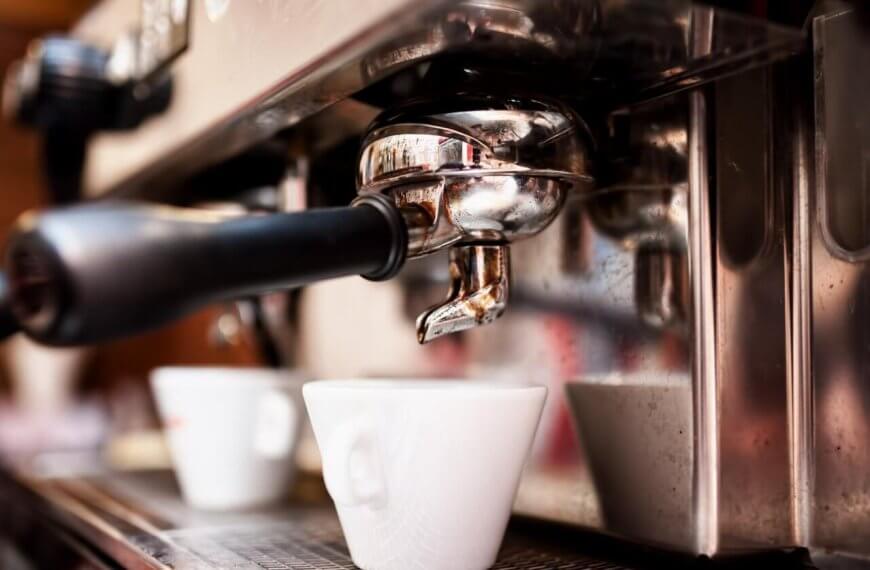 Close-up of a professional espresso machine brewing coffee into a white ceramic cup. The portafilter and spout are shiny and metallic, fixed securely in place. The cup sits on a metal drip tray, catching dark espresso as it flows from the machine. Experience this at one of the best restaurants in Gateway Mall, Quezon City.
