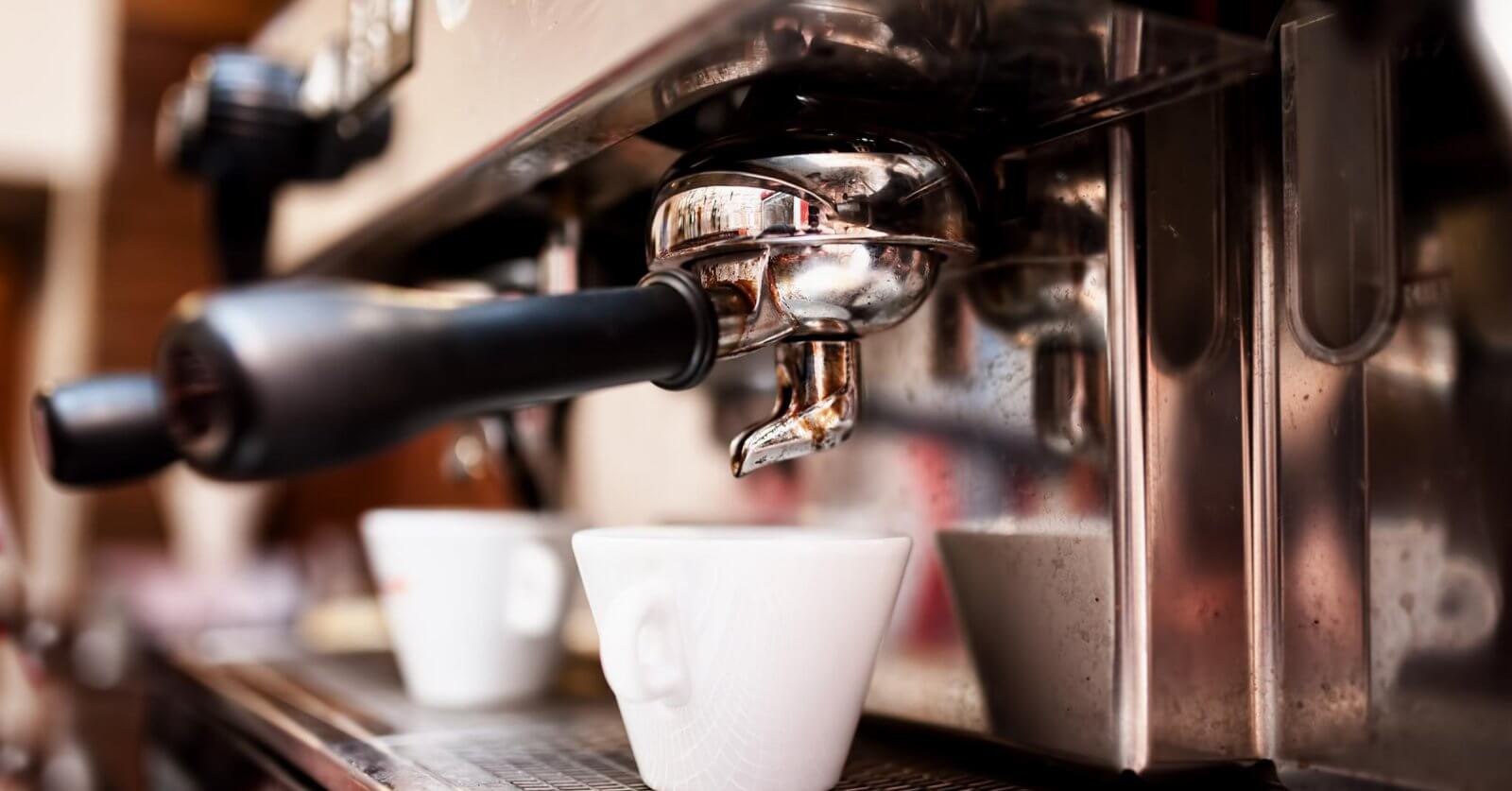 Close-up of a professional espresso machine brewing coffee into a white ceramic cup. The portafilter and spout are shiny and metallic, fixed securely in place. The cup sits on a metal drip tray, catching dark espresso as it flows from the machine. Experience this at one of the best restaurants in Gateway Mall, Quezon City.