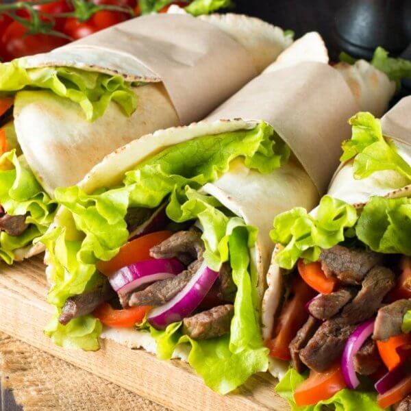 Three wraps filled with seasoned beef strips, fresh lettuce, sliced red onions, and red bell peppers are displayed on a wooden board at one of Makati's renowned Greenbelt Mall restaurants. The wraps are partially wrapped in parchment paper. In the background, there is a bowl of red sauce, cherry tomatoes, and a whole red chili pepper.