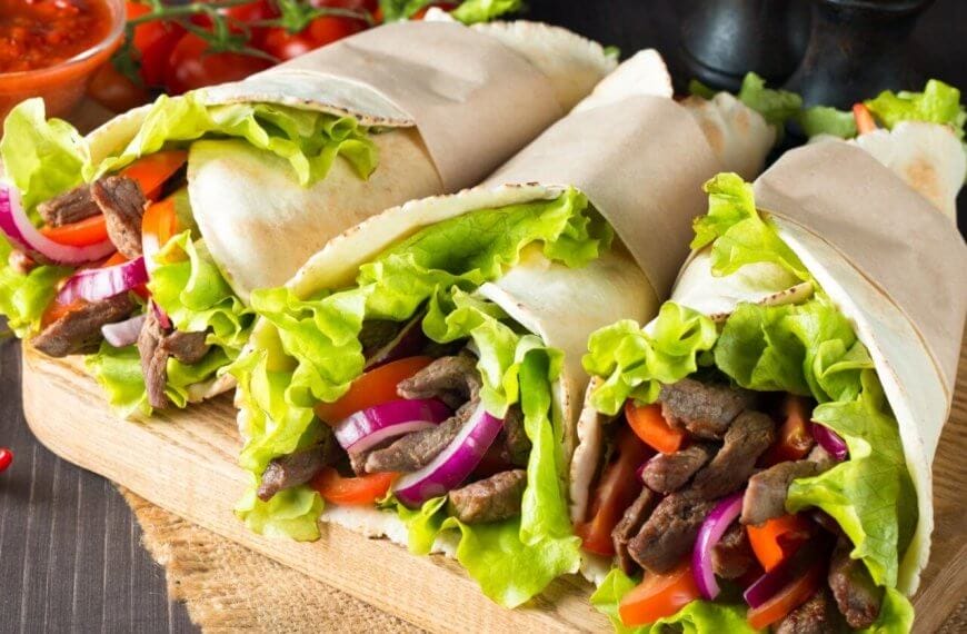 Three wraps filled with seasoned beef strips, fresh lettuce, sliced red onions, and red bell peppers are displayed on a wooden board at one of Makati's renowned Greenbelt Mall restaurants. The wraps are partially wrapped in parchment paper. In the background, there is a bowl of red sauce, cherry tomatoes, and a whole red chili pepper.