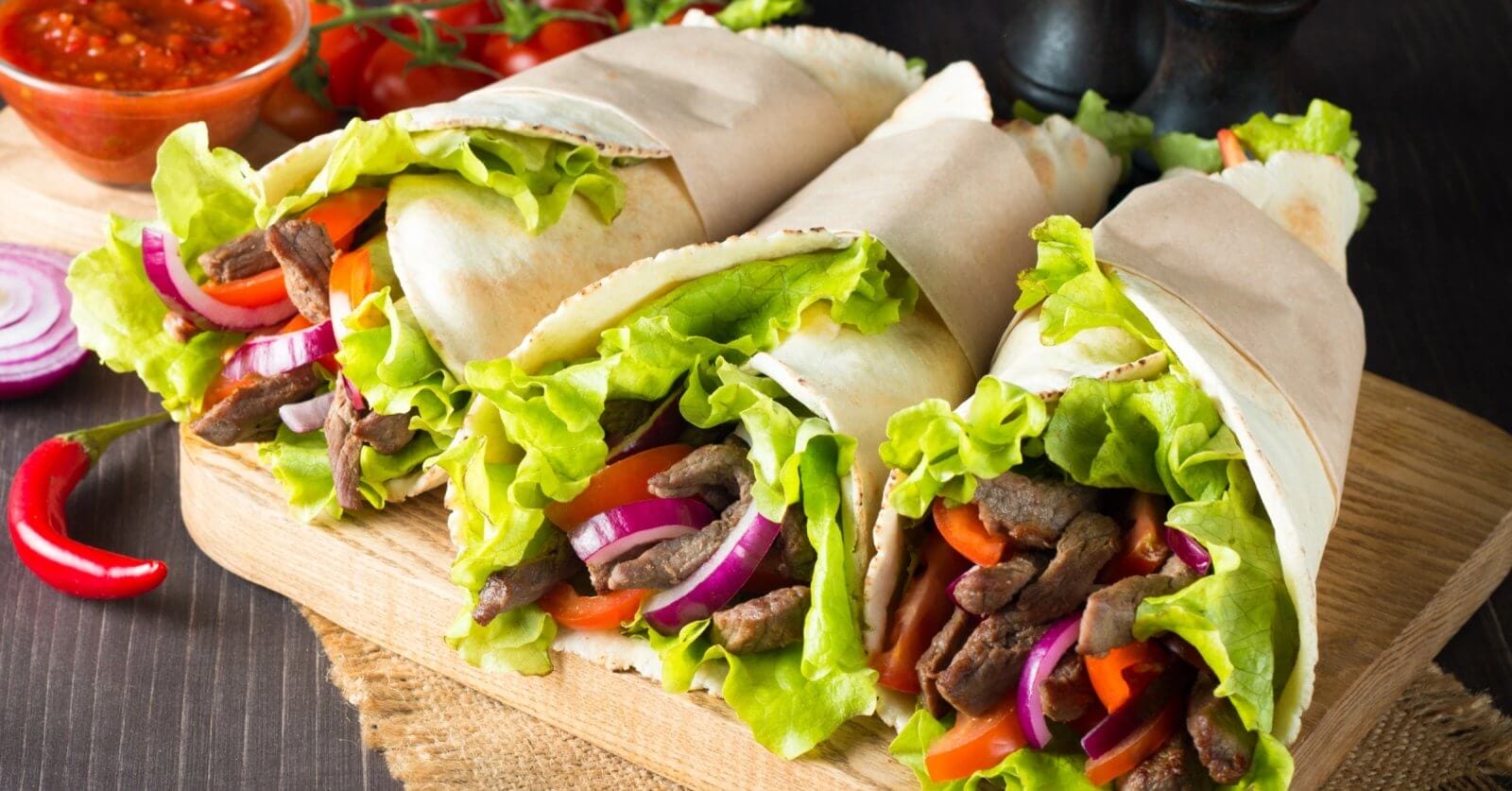Three wraps filled with seasoned beef strips, fresh lettuce, sliced red onions, and red bell peppers are displayed on a wooden board at one of Makati's renowned Greenbelt Mall restaurants. The wraps are partially wrapped in parchment paper. In the background, there is a bowl of red sauce, cherry tomatoes, and a whole red chili pepper.