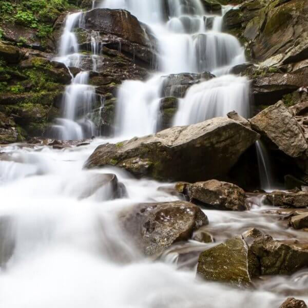 A cascading waterfall flows over multiple rocky tiers surrounded by lush green foliage, a highlight among Iligan's tourist attractions. The water appears smooth and silky, contrasting with the rugged, moss-covered rocks. The scene conveys a tranquil and refreshing natural environment.