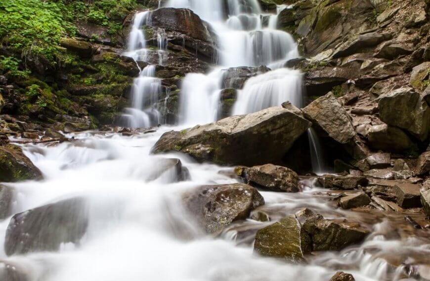 A cascading waterfall flows over multiple rocky tiers surrounded by lush green foliage, a highlight among Iligan's tourist attractions. The water appears smooth and silky, contrasting with the rugged, moss-covered rocks. The scene conveys a tranquil and refreshing natural environment.