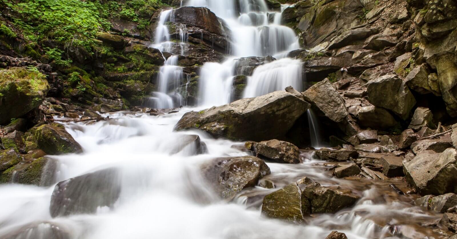 A cascading waterfall flows over multiple rocky tiers surrounded by lush green foliage, a highlight among Iligan's tourist attractions. The water appears smooth and silky, contrasting with the rugged, moss-covered rocks. The scene conveys a tranquil and refreshing natural environment.