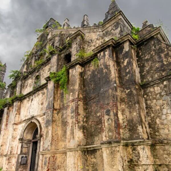 An ancient, weathered church facade stands against a dark, cloudy sky in the Ilocos Region. The stone structure features arches, ornate carvings, and an adjacent bell tower. Vines and plants grow out of the stone, emphasizing its aged and abandoned appearance. The scene exudes a historical and dramatic atmosphere.
