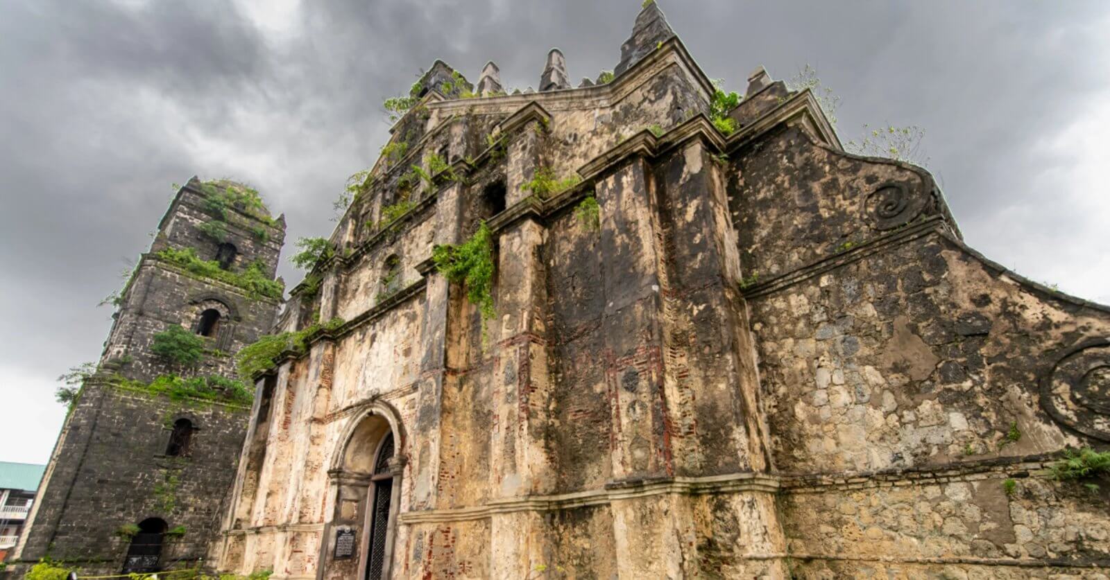 An ancient, weathered church facade stands against a dark, cloudy sky in the Ilocos Region. The stone structure features arches, ornate carvings, and an adjacent bell tower. Vines and plants grow out of the stone, emphasizing its aged and abandoned appearance. The scene exudes a historical and dramatic atmosphere.