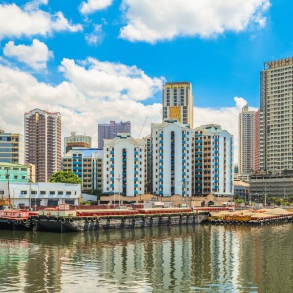 A vibrant cityscape in Manila shows a mix of modern and older buildings along a waterfront. Several boats and barges are docked, reflecting on the calm water. The sky is bright with scattered clouds, adding to the lively atmosphere of this urban harbor scene, one of the city's popular tourist attractions.