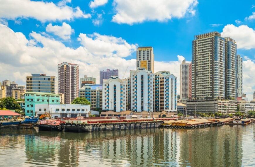 A vibrant cityscape in Manila shows a mix of modern and older buildings along a waterfront. Several boats and barges are docked, reflecting on the calm water. The sky is bright with scattered clouds, adding to the lively atmosphere of this urban harbor scene, one of the city's popular tourist attractions.