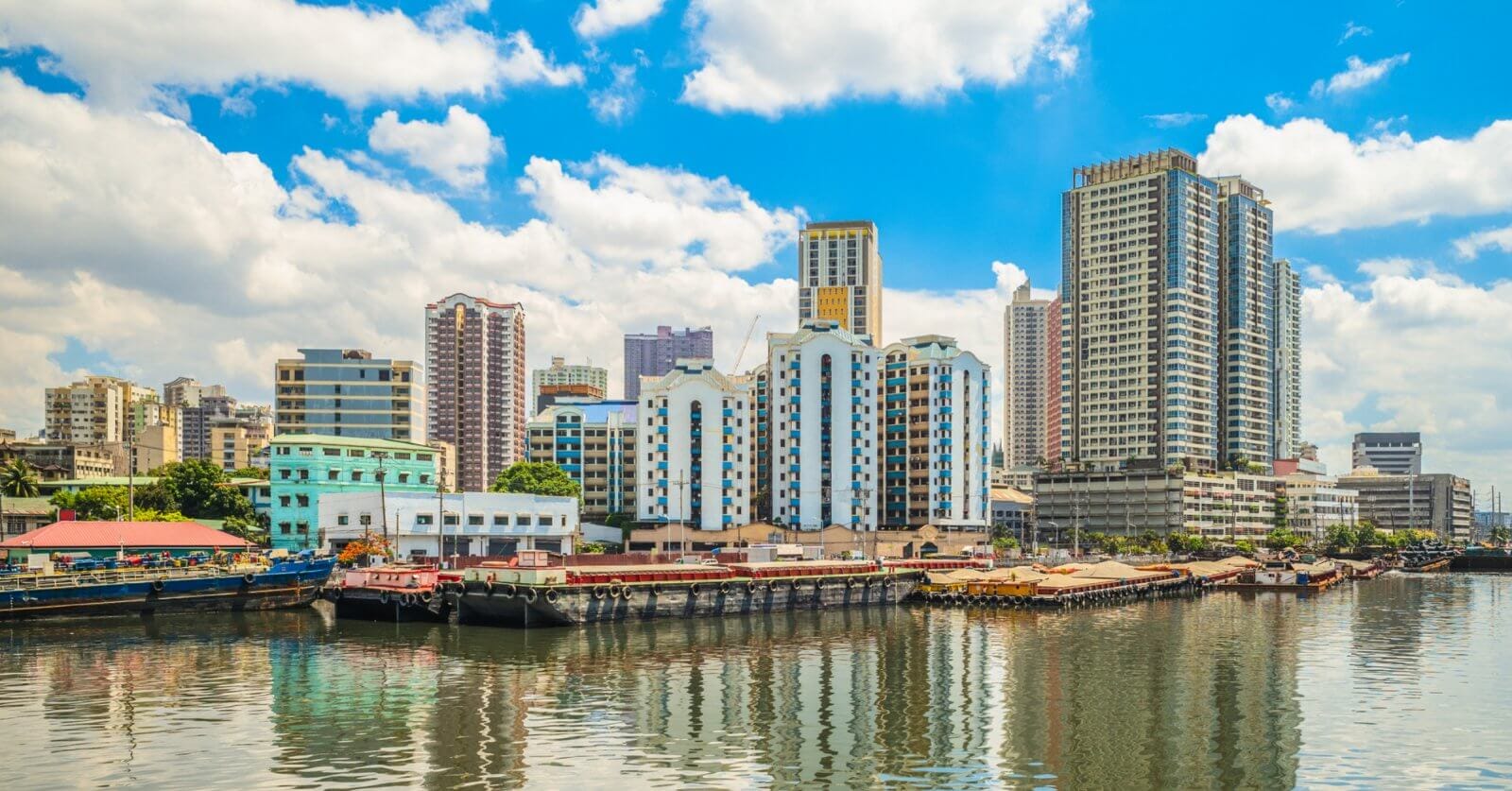 A vibrant cityscape in Manila shows a mix of modern and older buildings along a waterfront. Several boats and barges are docked, reflecting on the calm water. The sky is bright with scattered clouds, adding to the lively atmosphere of this urban harbor scene, one of the city's popular tourist attractions.