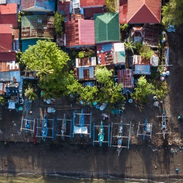 Aerial view of a coastal village in Region 4B, featuring numerous small, colorful houses with tin roofs. Many houses are surrounded by trees. Fishing boats line the dark, sandy shoreline, casting long shadows. The beach rests in the foreground, transitioning into the bluish water at the bottom of the image.