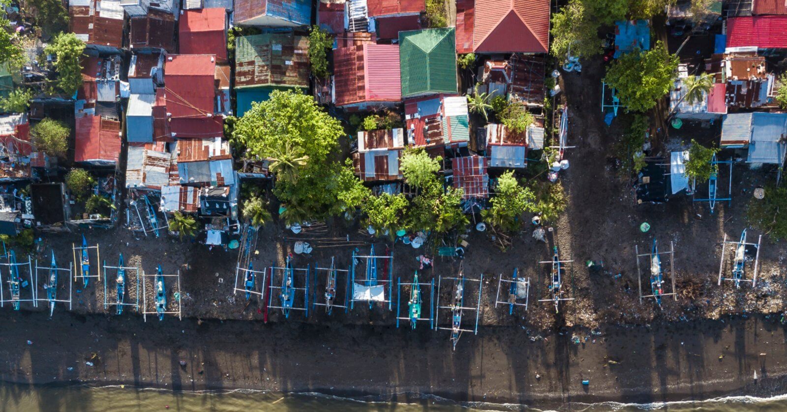 Aerial view of a coastal village in Region 4B, featuring numerous small, colorful houses with tin roofs. Many houses are surrounded by trees. Fishing boats line the dark, sandy shoreline, casting long shadows. The beach rests in the foreground, transitioning into the bluish water at the bottom of the image.