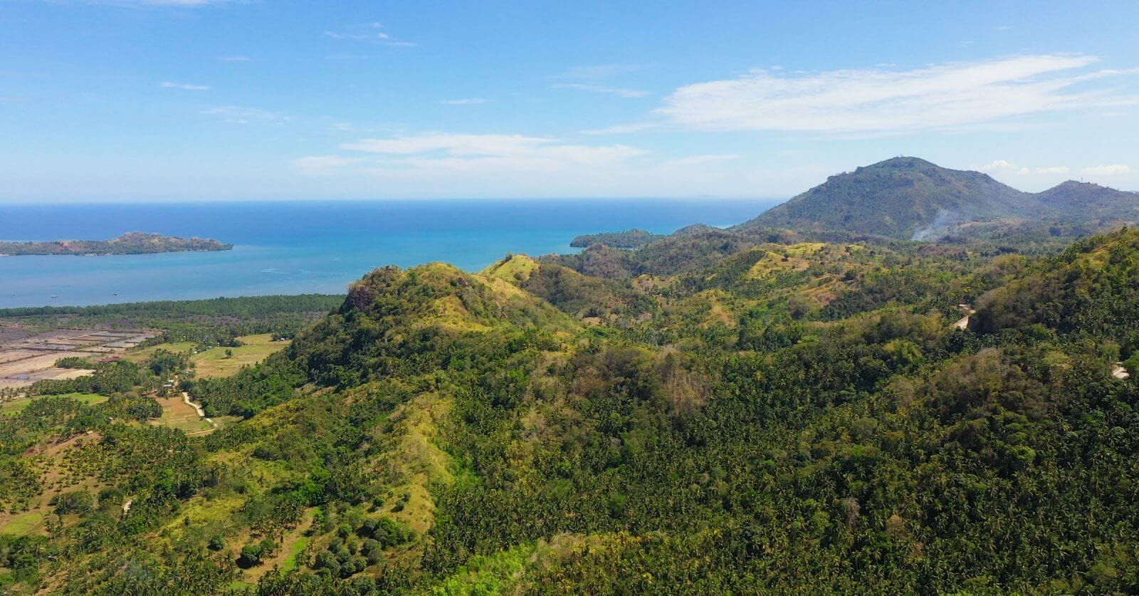 Aerial view of Northern Mindanao's lush, green hilly landscape stretching towards a calm, bright blue sea under a clear sky. Dense forests cover the hills, with patches of fields and sparse trees. In the distance, low-lying lands extend to the shoreline, with mountains visible on the horizon.