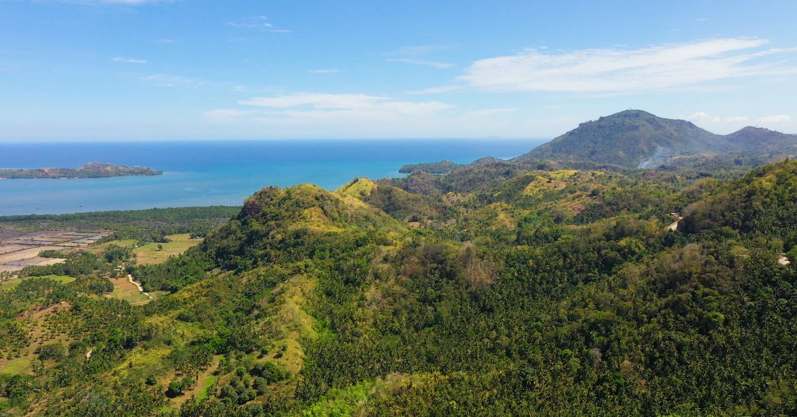 Aerial view of Northern Mindanao's lush, green hilly landscape stretching towards a calm, bright blue sea under a clear sky. Dense forests cover the hills, with patches of fields and sparse trees. In the distance, low-lying lands extend to the shoreline, with mountains visible on the horizon.