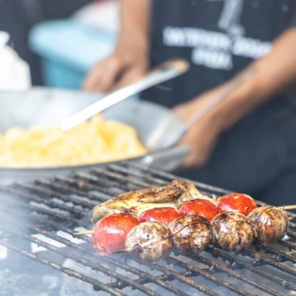 A person grills skewers of mushrooms and cherry tomatoes over a flame. In the background, another person is cooking a dish in a large wok. Both individuals are partially out of focus, with the grilling food as the main subject. The lively scene seems to capture one of Quezon City's best outdoor market spots.