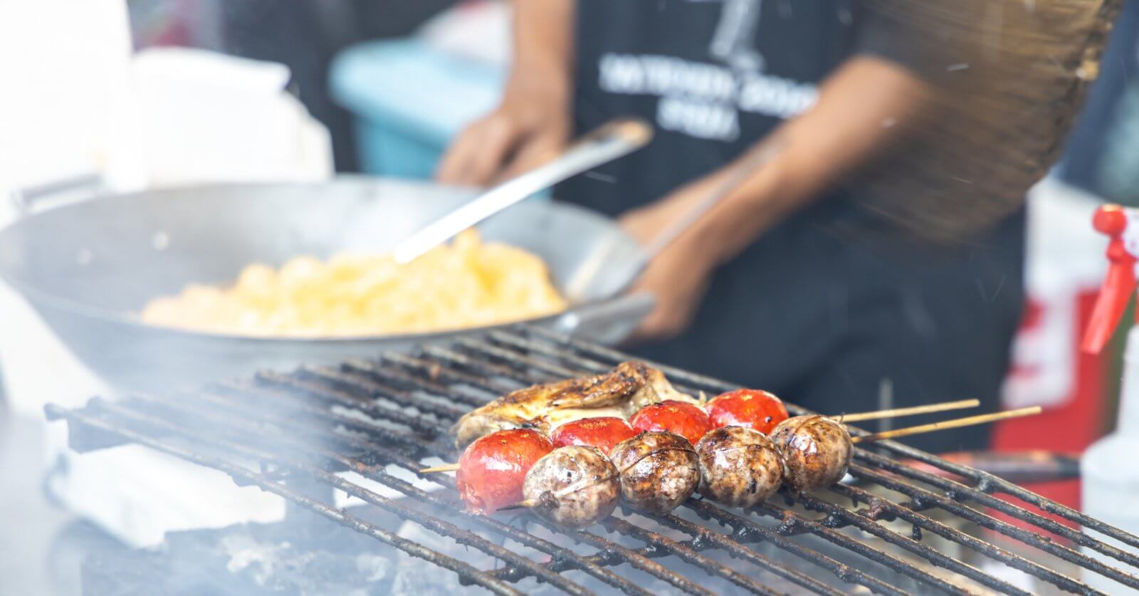 A person grills skewers of mushrooms and cherry tomatoes over a flame. In the background, another person is cooking a dish in a large wok. Both individuals are partially out of focus, with the grilling food as the main subject. The lively scene seems to capture one of Quezon City's best outdoor market spots.