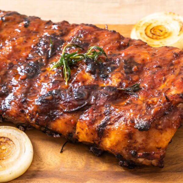 A slab of grilled barbecue ribs rests on a wooden cutting board, adorned with char marks and a coating of sauce. Garnishes of roasted cherry tomatoes, whole onion slices, and rosemary sprigs are arranged around the ribs, enhancing the visual appeal of the dish—perfect for dining at SM Fairview restaurants in Quezon City.