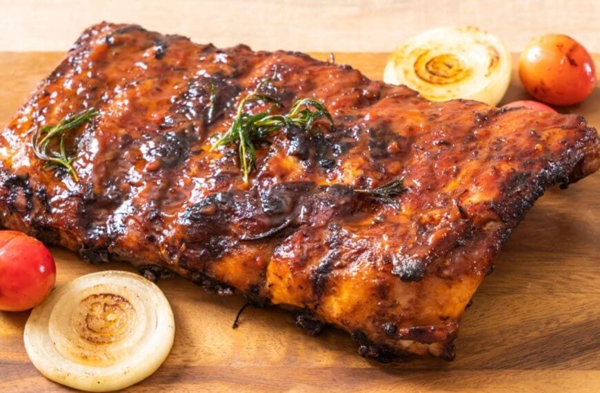 A slab of grilled barbecue ribs rests on a wooden cutting board, adorned with char marks and a coating of sauce. Garnishes of roasted cherry tomatoes, whole onion slices, and rosemary sprigs are arranged around the ribs, enhancing the visual appeal of the dish—perfect for dining at SM Fairview restaurants in Quezon City.