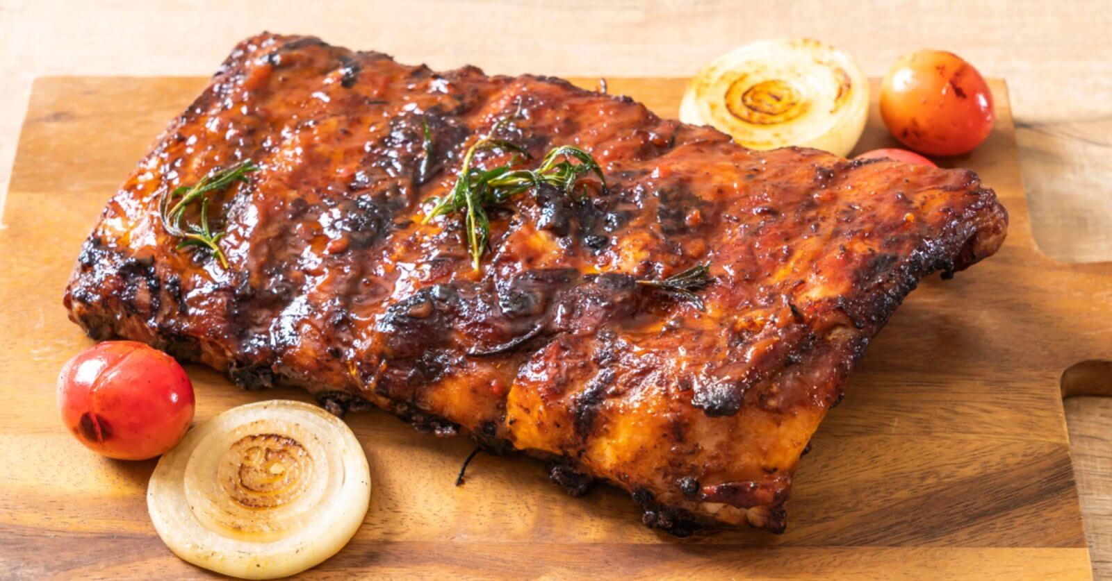 A slab of grilled barbecue ribs rests on a wooden cutting board, adorned with char marks and a coating of sauce. Garnishes of roasted cherry tomatoes, whole onion slices, and rosemary sprigs are arranged around the ribs, enhancing the visual appeal of the dish—perfect for dining at SM Fairview restaurants in Quezon City.