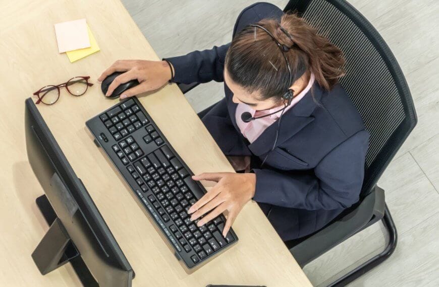 A person wearing a headset is seated at a desk using a computer keyboard and mouse, possibly working as one of the best virtual assistants. The individual is dressed in business attire. The desk has eyeglasses and sticky notes on it. The scene is viewed from an overhead perspective.