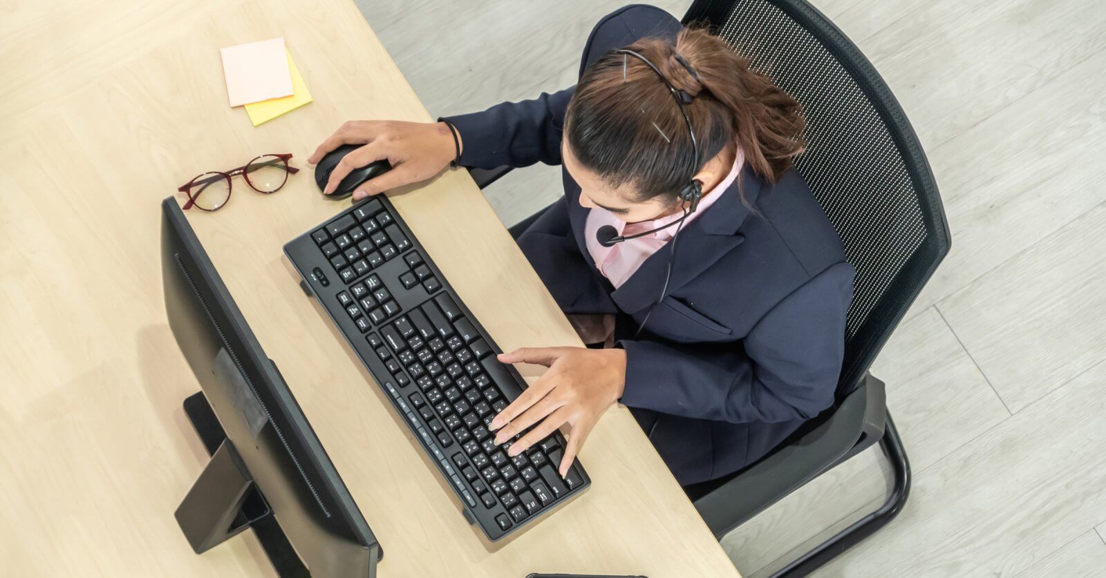 A person wearing a headset is seated at a desk using a computer keyboard and mouse, possibly working as one of the best virtual assistants. The individual is dressed in business attire. The desk has eyeglasses and sticky notes on it. The scene is viewed from an overhead perspective.