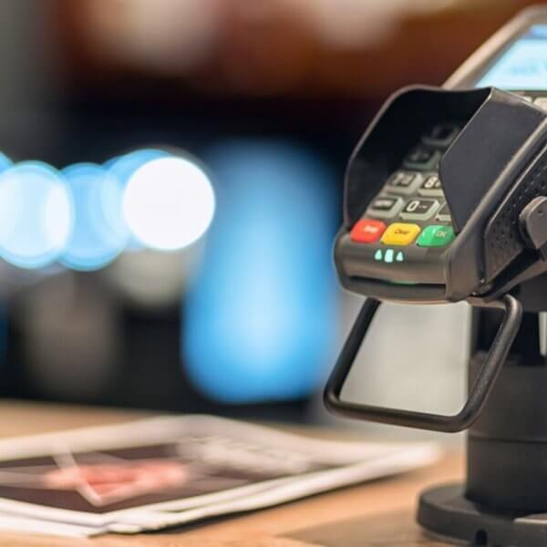 Close-up of a credit card terminal mounted on a stand at a checkout counter, supplied by Sulit Business Solutions. The terminal display is lit, showing some text. Nearby, blurred brochures and soft-focus lights create a cozy ambiance. The terminal features red, yellow, and green buttons.