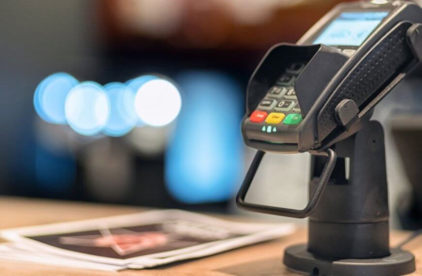 Close-up of a credit card terminal mounted on a stand at a checkout counter, supplied by Sulit Business Solutions. The terminal display is lit, showing some text. Nearby, blurred brochures and soft-focus lights create a cozy ambiance. The terminal features red, yellow, and green buttons.