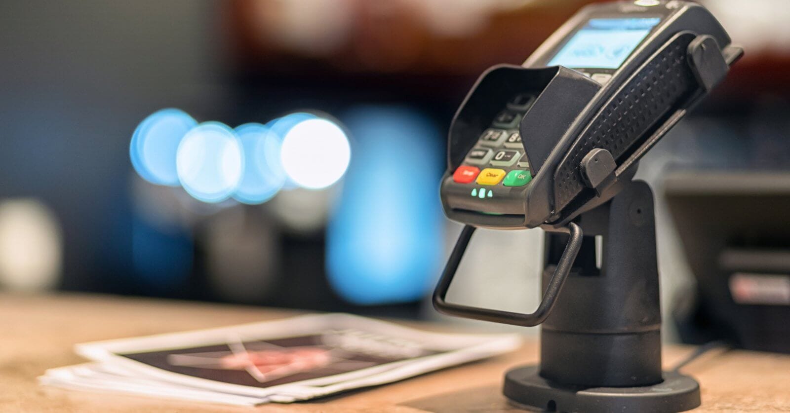 Close-up of a credit card terminal mounted on a stand at a checkout counter, supplied by Sulit Business Solutions. The terminal display is lit, showing some text. Nearby, blurred brochures and soft-focus lights create a cozy ambiance. The terminal features red, yellow, and green buttons.