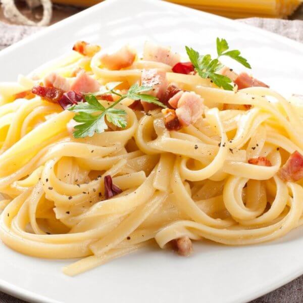 A plate of pasta carbonara on a white square dish from one of Bridgetowne's charming restaurants. The dish features fettuccine noodles coated in a creamy sauce, sprinkled with bacon pieces and black pepper, and garnished with fresh parsley. Set on a brown cloth with a rustic wooden background.