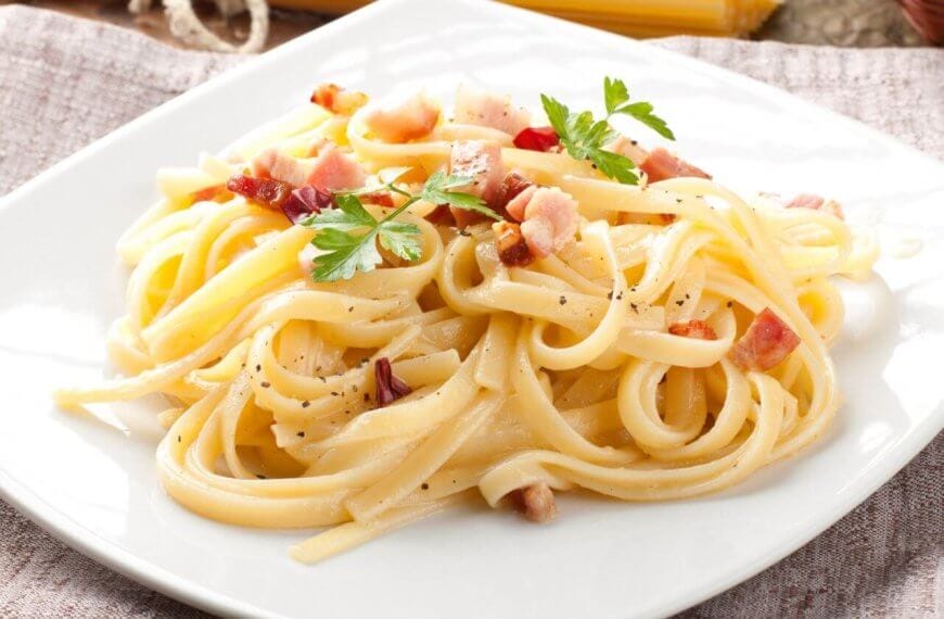 A plate of pasta carbonara on a white square dish from one of Bridgetowne's charming restaurants. The dish features fettuccine noodles coated in a creamy sauce, sprinkled with bacon pieces and black pepper, and garnished with fresh parsley. Set on a brown cloth with a rustic wooden background.