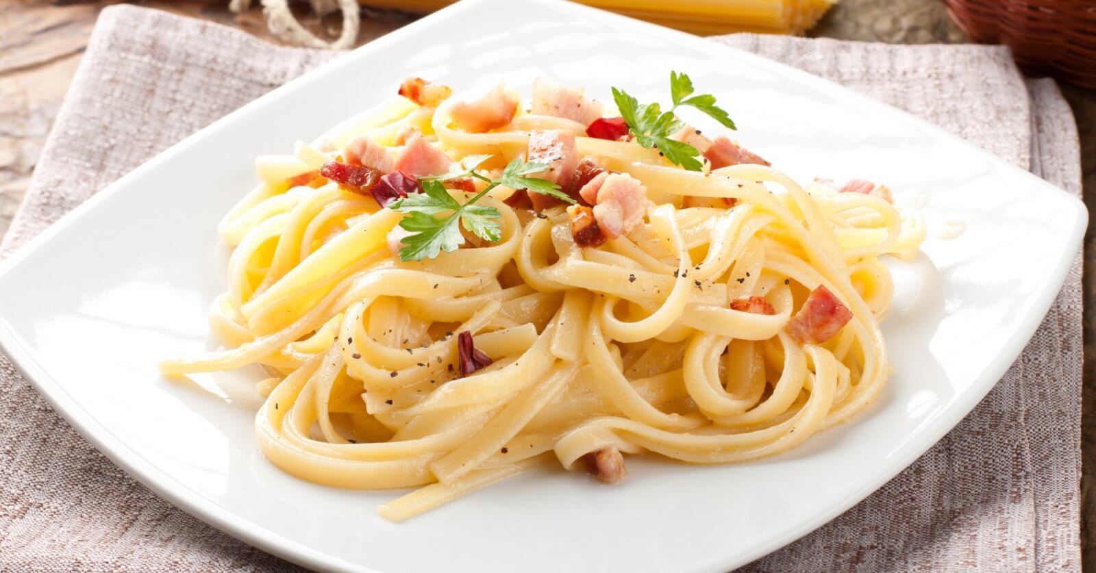 A plate of pasta carbonara on a white square dish from one of Bridgetowne's charming restaurants. The dish features fettuccine noodles coated in a creamy sauce, sprinkled with bacon pieces and black pepper, and garnished with fresh parsley. Set on a brown cloth with a rustic wooden background.