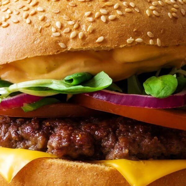 A close-up of one of Metro Manila's best burgers: a sesame seed bun enveloping layers of melted cheese, a juicy beef patty, sliced red onions, a tomato slice, fresh spinach leaves, and a generous layer of creamy sauce. The slightly blurred background accentuates the burger's vibrant textures and colors.
