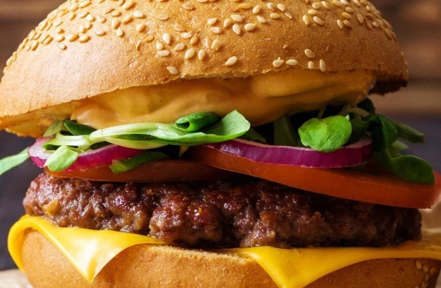 A close-up of one of Metro Manila's best burgers: a sesame seed bun enveloping layers of melted cheese, a juicy beef patty, sliced red onions, a tomato slice, fresh spinach leaves, and a generous layer of creamy sauce. The slightly blurred background accentuates the burger's vibrant textures and colors.