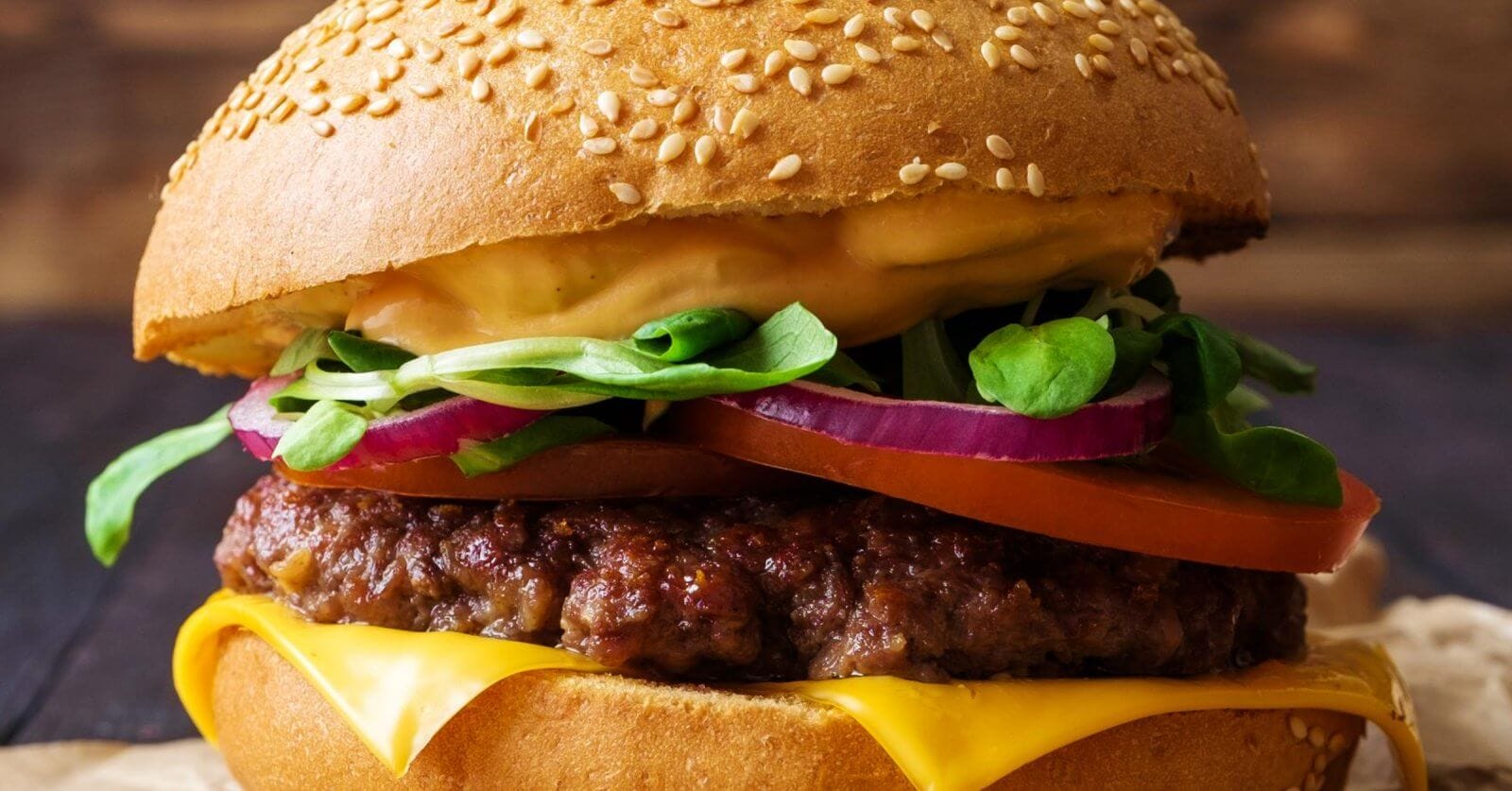 A close-up of one of Metro Manila's best burgers: a sesame seed bun enveloping layers of melted cheese, a juicy beef patty, sliced red onions, a tomato slice, fresh spinach leaves, and a generous layer of creamy sauce. The slightly blurred background accentuates the burger's vibrant textures and colors.