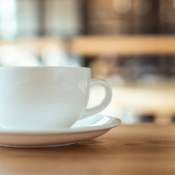 A white coffee cup rests on a matching saucer on a wooden table, embodying the essence of Metro Manila's best coffee. The blurred background, with warm tones and hints of light, suggests a cozy cafe atmosphere, evoking a sense of calm and relaxation.