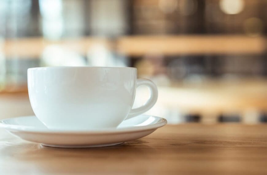 A white coffee cup rests on a matching saucer on a wooden table, embodying the essence of Metro Manila's best coffee. The blurred background, with warm tones and hints of light, suggests a cozy cafe atmosphere, evoking a sense of calm and relaxation.