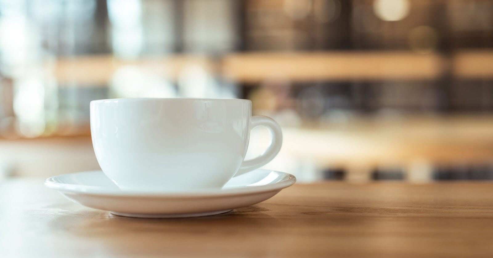 A white coffee cup rests on a matching saucer on a wooden table, embodying the essence of Metro Manila's best coffee. The blurred background, with warm tones and hints of light, suggests a cozy cafe atmosphere, evoking a sense of calm and relaxation.