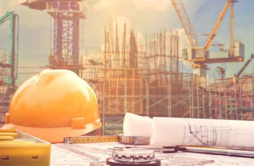 A construction site with cranes and scaffolding rises under Manila's blue sky. In the foreground, a yellow hard hat, rolled blueprints, a measuring tape, and a compass rest on a table, symbolizing architectural planning backed by construction supply companies known for quality and reliability.