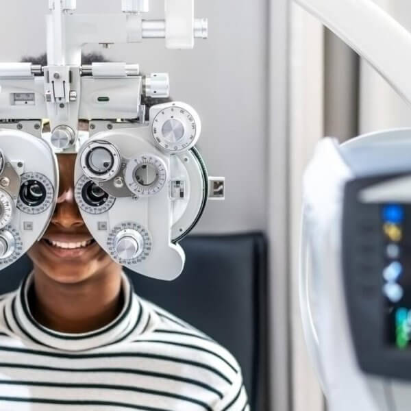 In one of Metro Manila's best eye clinics, a person in a striped shirt sits behind a phoropter, undergoing an eye examination. The focus is on the device with its various dials and lenses, while a blurred vision-testing machine looms in the foreground. The scene is set in a bright, clinical room.