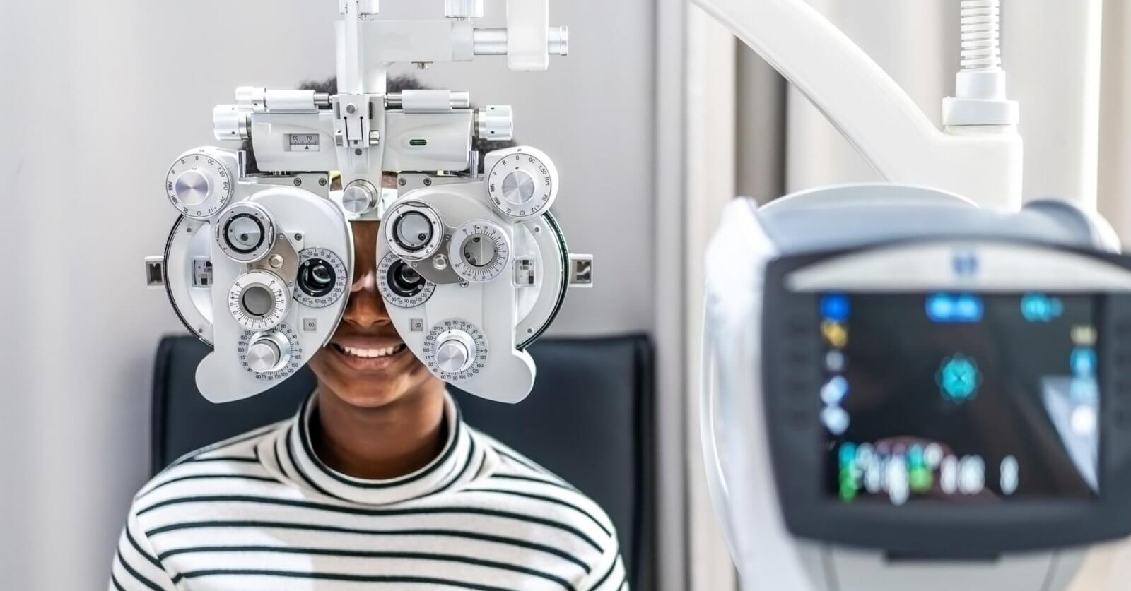In one of Metro Manila's best eye clinics, a person in a striped shirt sits behind a phoropter, undergoing an eye examination. The focus is on the device with its various dials and lenses, while a blurred vision-testing machine looms in the foreground. The scene is set in a bright, clinical room.