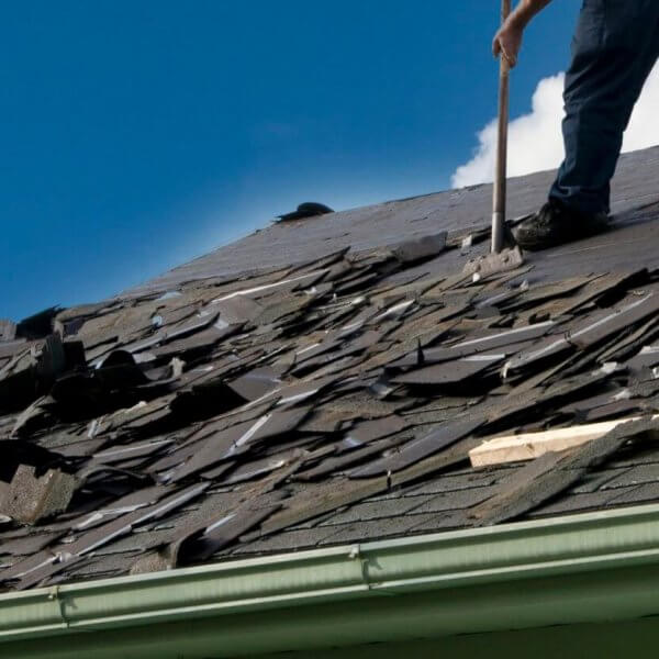 A person is removing old shingles from a sloped roof with a tool sourced from premium roofing suppliers. The roof, with its damaged and scattered shingles, needs attention. Wearing jeans, the person stands against a clear blue sky with some clouds, while a green gutter lines the edge of the roof.