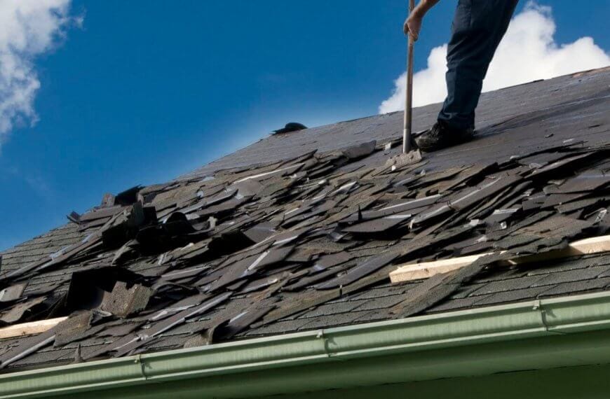 A person is removing old shingles from a sloped roof with a tool sourced from premium roofing suppliers. The roof, with its damaged and scattered shingles, needs attention. Wearing jeans, the person stands against a clear blue sky with some clouds, while a green gutter lines the edge of the roof.