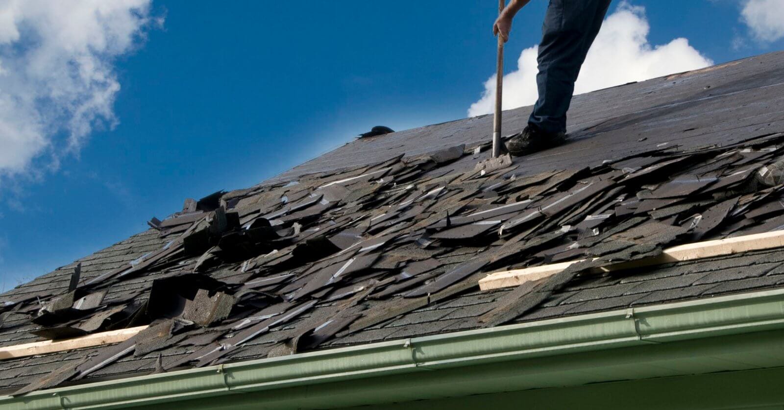 A person is removing old shingles from a sloped roof with a tool sourced from premium roofing suppliers. The roof, with its damaged and scattered shingles, needs attention. Wearing jeans, the person stands against a clear blue sky with some clouds, while a green gutter lines the edge of the roof.