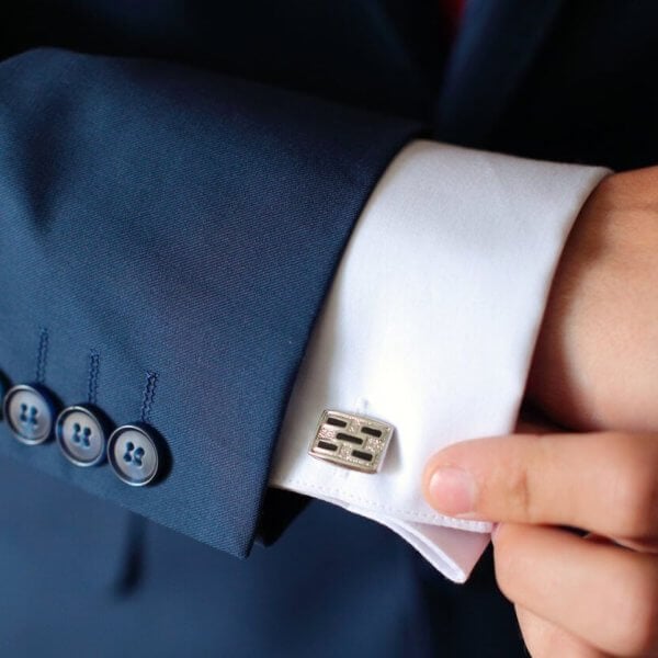 A person expertly adjusts a metallic, rectangular cufflink on a fitted white dress shirt. The tailored sleeve, part of a dark blue suit with four blue buttons, exudes elegance. The setting is shadowed and elusive, reminiscent of Metro Manila's sophisticated style.